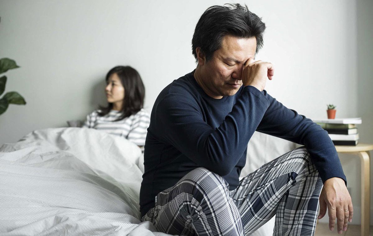 Asian man sitting on edge of bed, looking irritated, while wife is looking away.